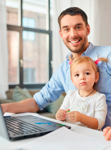 Junger Vater mit Tochter auf dem Schoss sitzt zu Hause am Laptop und arbeitet in seiner Freizeit